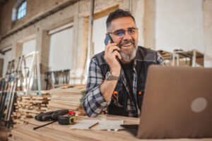 Carpenter in wood workshop using laptop and working on project, online shopping or banking.