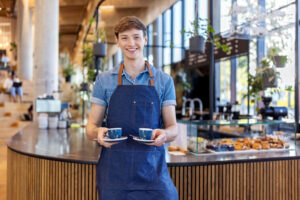 Smiling young male barista serving two cups of coffee at office cafeteria. Young man giving coffee to customer at cafe.