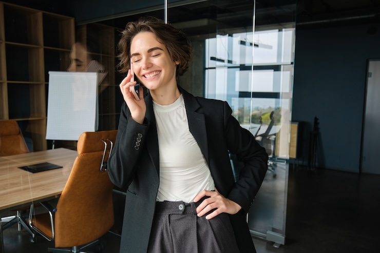 Image of successful businesswoman having a telephone conversation in an office with glass wall, smiling and looking satisfied with negotiations.